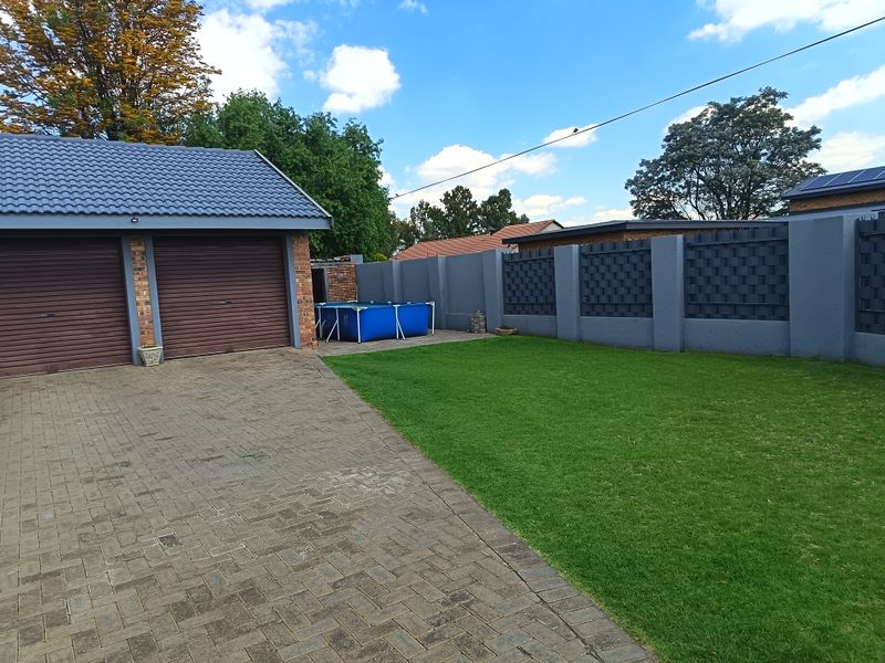A well-maintained rear garden area featuring a paved driveway, double garage with roller doors, and a secure perimeter boundary wall.