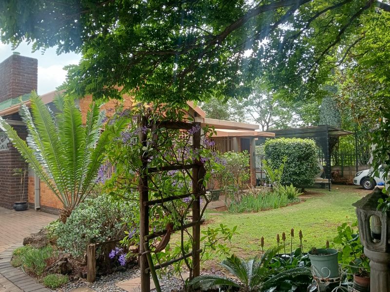 A view of the rear garden featuring a brick facade, paved patio area, and established landscaping under mature trees.