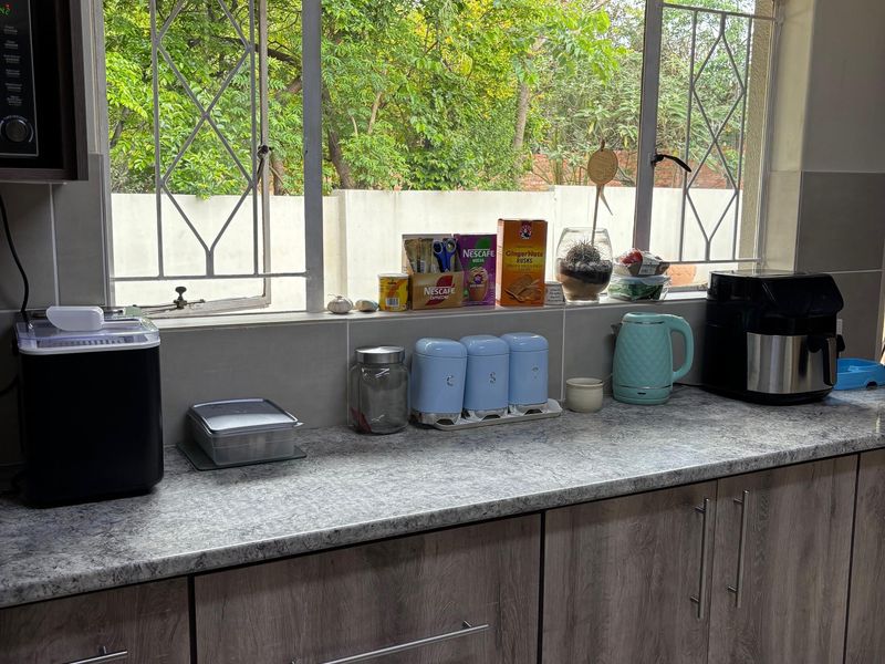 A functional kitchen area featuring modern wood-look cabinetry, a durable laminate countertop, and a large window with security bars overlooking the garden.