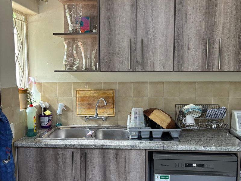 A functional kitchen area featuring wood-look laminate cabinetry, a double basin sink with mixer tap, tiled backsplash, and an integrated dishwasher.