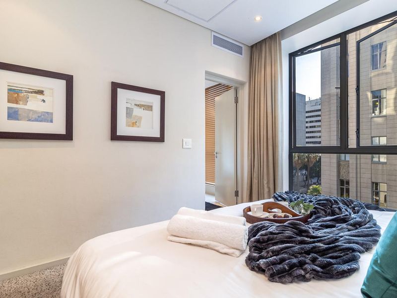 A well-maintained bedroom featuring neutral walls, carpeted flooring, recessed lighting, ducted air conditioning, and a large window providing natural light and urban views.