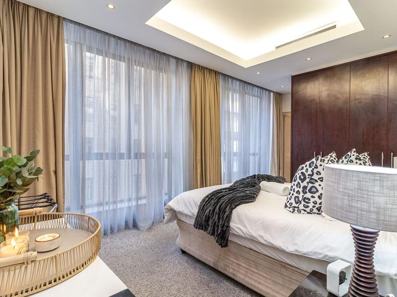 A well-appointed bedroom featuring a tray ceiling with recessed lighting, built-in dark wood wardrobes, and large windows with floor-to-ceiling curtains.