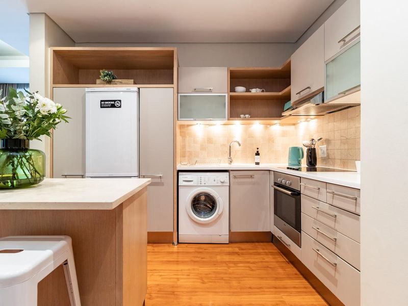 Modern kitchen featuring light-toned cabinetry, stone countertops, integrated appliances, under-cabinet lighting, and a functional under-counter washing machine.
