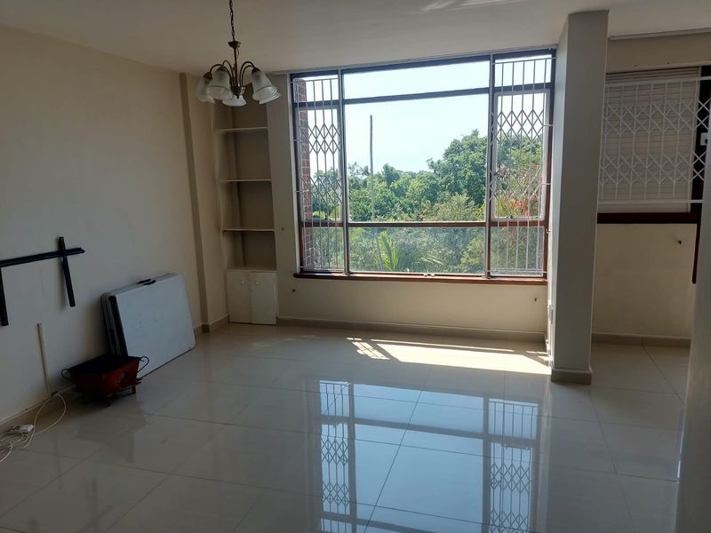 A bright, empty living room featuring large windows with security gates, built-in shelving, and polished tiled flooring, offering a pleasant view of greenery.