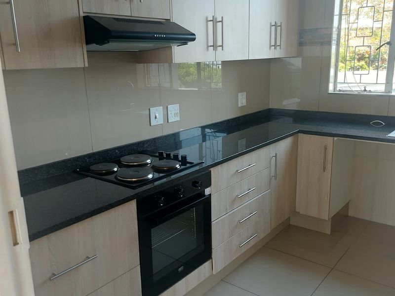 Modern kitchen featuring light wood-finish cabinetry, dark granite countertops, integrated electric hob and oven, and tiled flooring with natural light.