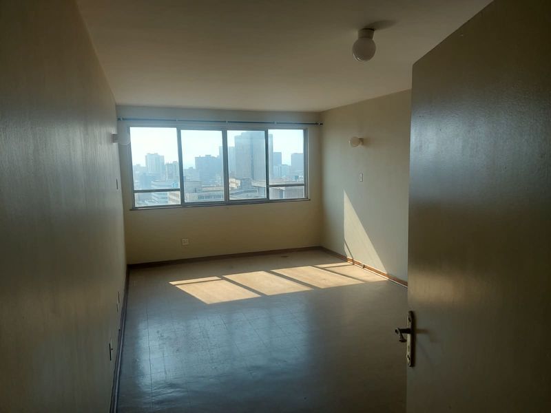 An empty bedroom featuring large windows with city views, vinyl flooring, and wall-mounted lighting, offering a blank canvas for renovation.