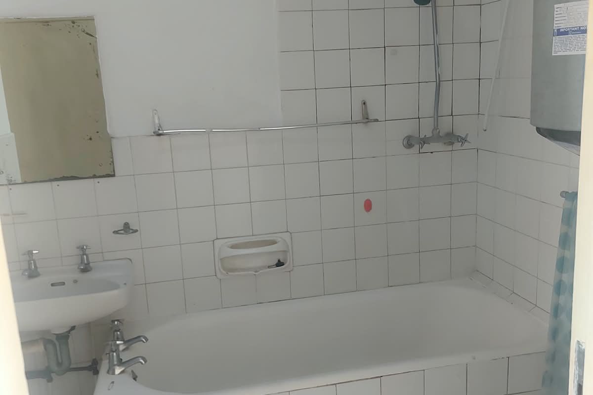A dated bathroom featuring a tiled bathtub, wall-mounted basin, and exposed water heater, requiring renovation to modernize the space.