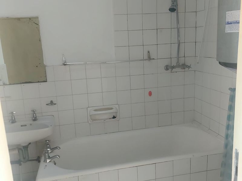 A dated bathroom featuring a tiled bathtub, wall-mounted basin, and exposed water heater, requiring renovation to modernize the space.