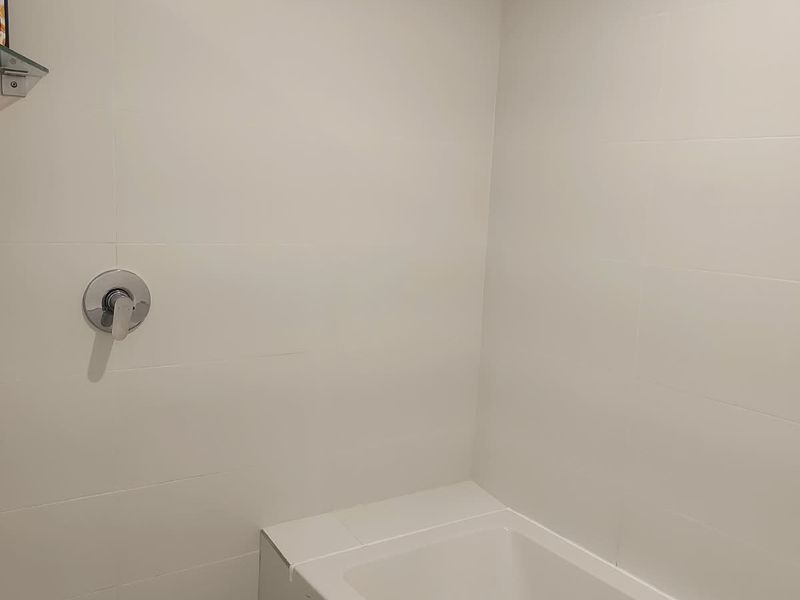A clean, minimalist bathroom featuring a built-in bathtub with a shower mixer and overhead shower head, finished with white wall tiles.