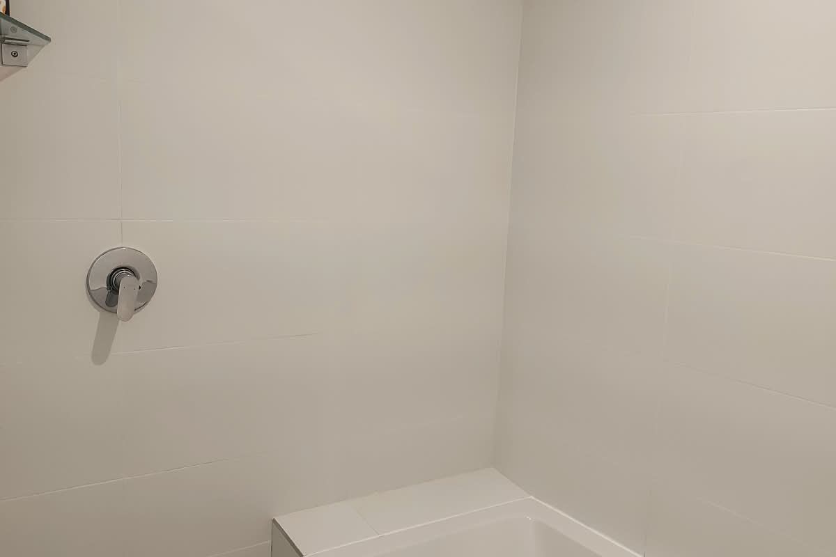 A clean, minimalist bathroom featuring a built-in bathtub with a shower mixer and overhead shower head, finished with white wall tiles.