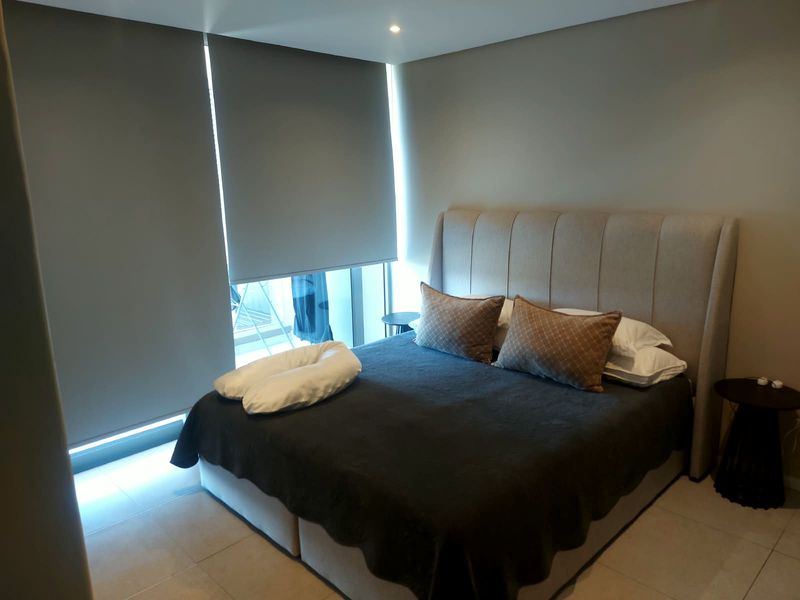A modern bedroom featuring neutral tiled flooring, recessed downlighting, and large roller blinds providing privacy and light control.