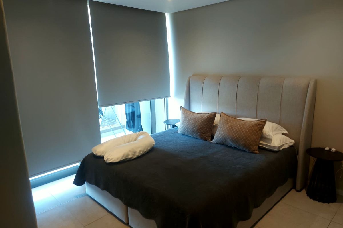 A modern bedroom featuring neutral tiled flooring, recessed downlighting, and large roller blinds providing privacy and light control.