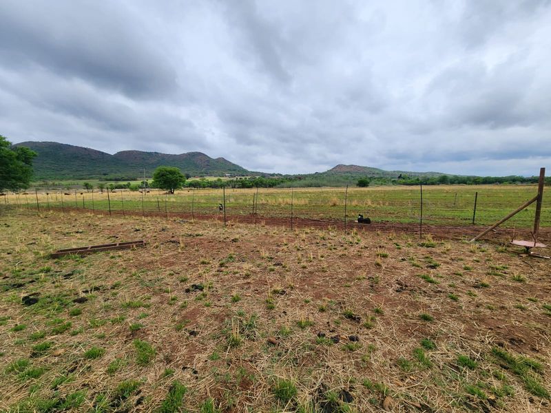 Expansive view of open farm pasture land enclosed by wire fencing with scenic mountain ranges in the background under a cloudy sky.