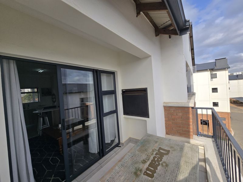 A covered balcony featuring a built-in braai, tiled flooring, and a metal balustrade, accessible via large sliding glass doors from the interior living space.