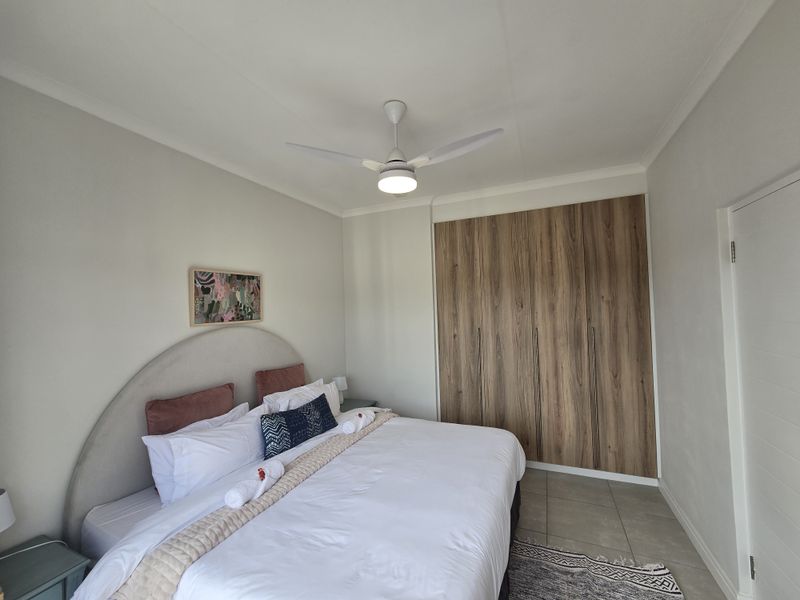 A well-maintained bedroom featuring modern wood-finish built-in cupboards, a ceiling fan, and neutral tiled flooring.