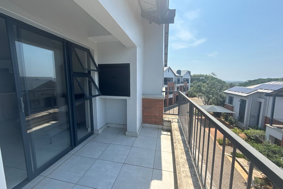 A spacious covered balcony featuring tiled flooring, a built-in braai, and a metal balustrade, offering an open view of the surrounding apartment complex.