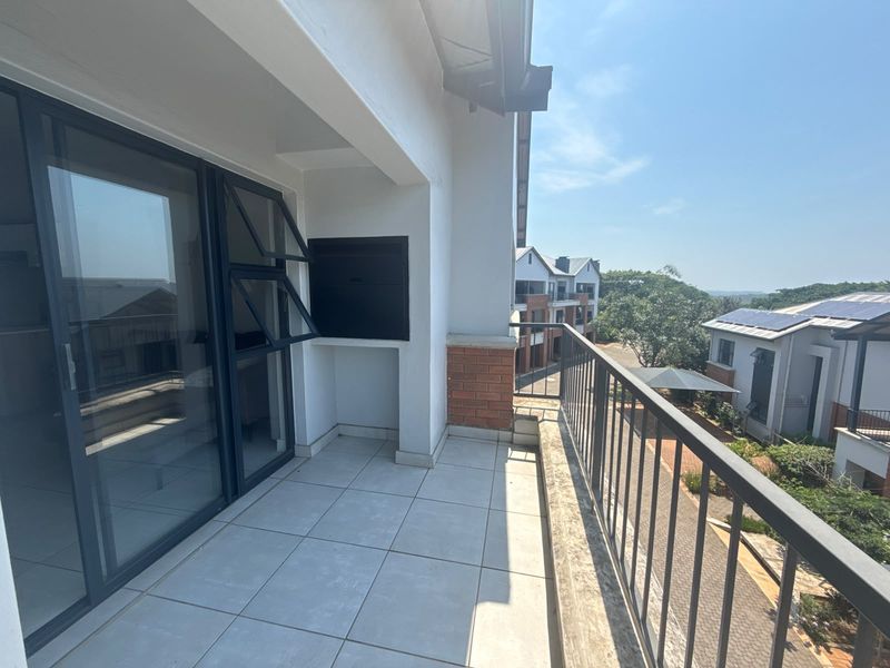 A spacious covered balcony featuring tiled flooring, a built-in braai, and a metal balustrade, offering an open view of the surrounding apartment complex.