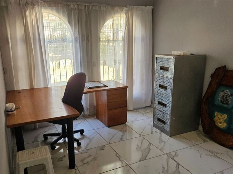 A functional home office space featuring large arched windows with security bars and modern marble-look tiled flooring.