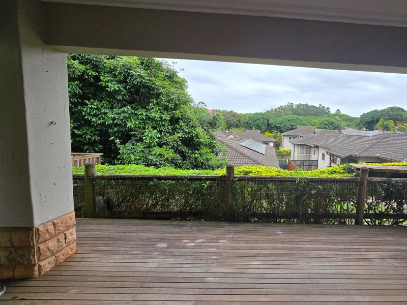 A covered outdoor patio featuring timber decking and stone-clad pillars, offering an elevated view of the surrounding neighborhood and lush greenery.