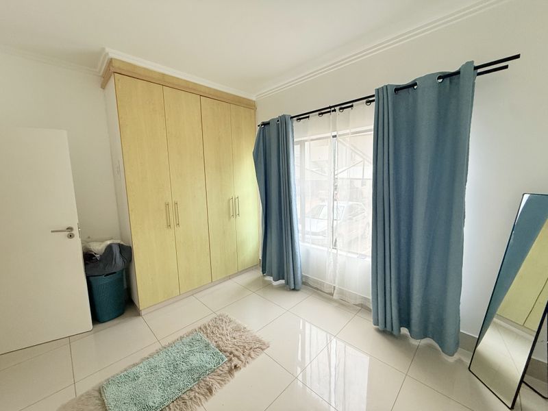 A bright bedroom featuring light wood built-in cupboards, neutral tiled flooring, and large windows with curtain fixtures.