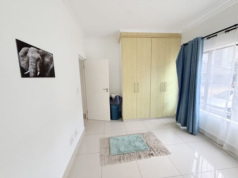 A bright bedroom featuring neutral tiled flooring, built-in wooden cupboards, and a large window with curtain fixtures.