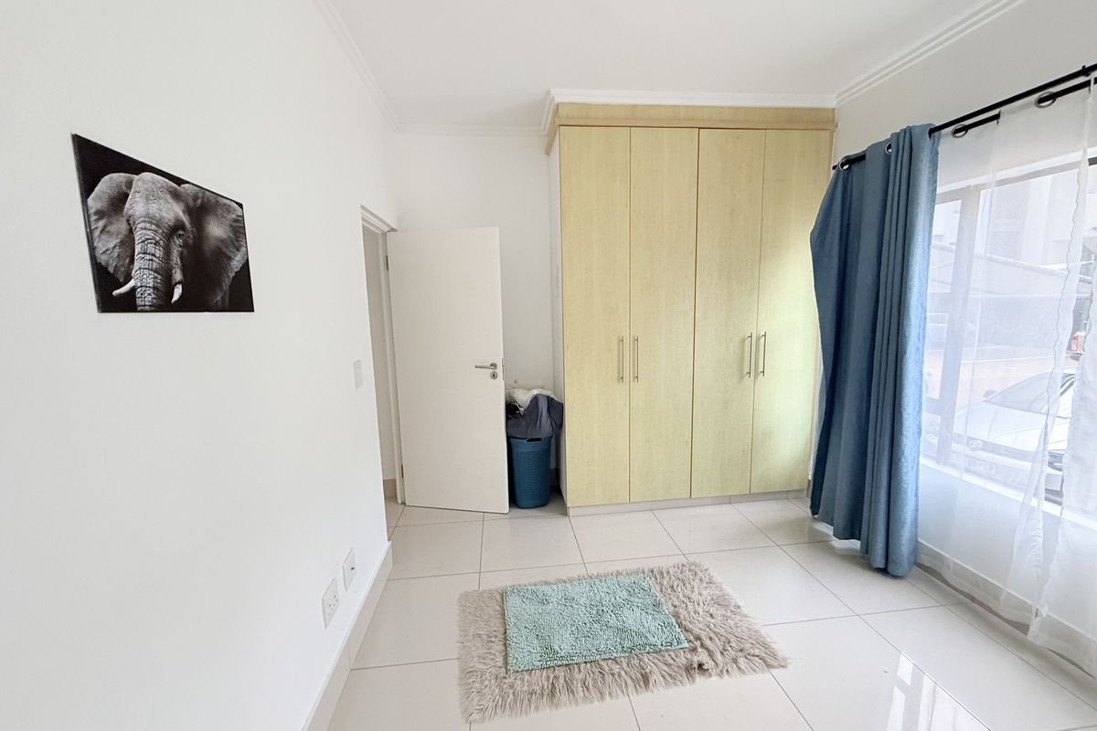 A bright bedroom featuring neutral tiled flooring, built-in wooden cupboards, and a large window with curtain fixtures.