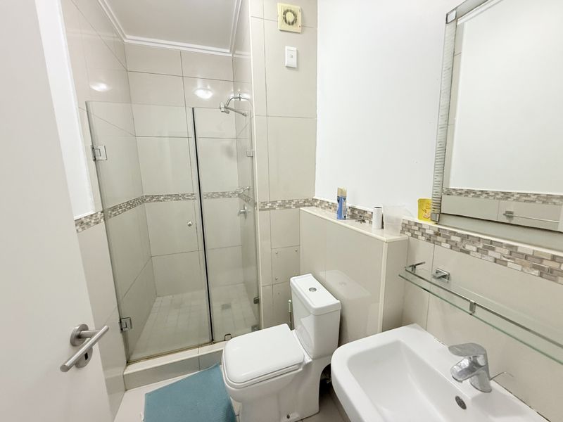 A clean and functional bathroom featuring a glass-enclosed shower, pedestal basin, and neutral wall tiling with a decorative mosaic border.