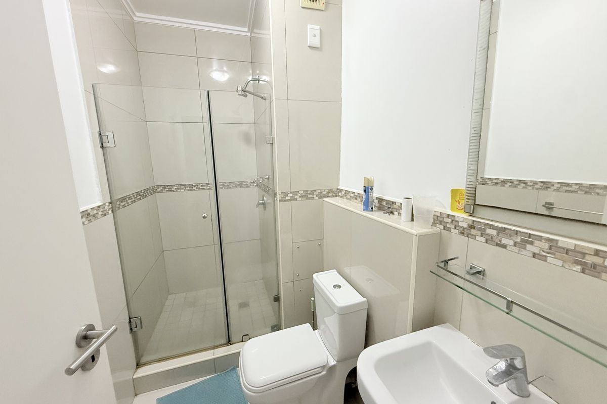 A clean and functional bathroom featuring a glass-enclosed shower, pedestal basin, and neutral wall tiling with a decorative mosaic border.