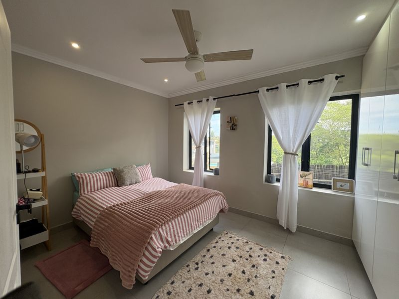A well-maintained bedroom featuring tiled flooring, built-in cupboards, a ceiling fan, and large black-framed windows allowing for natural light.