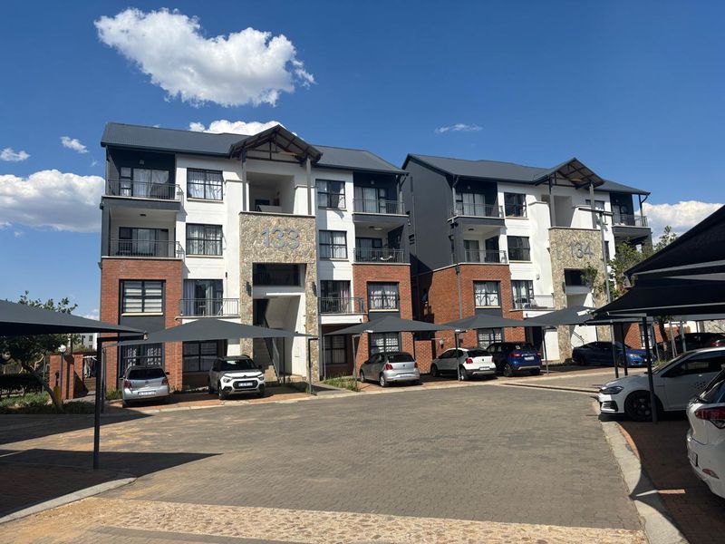 Front elevation of a multi-story apartment complex featuring brick and stone facade finishes, private balconies, and dedicated paved parking with shade cloth structures.