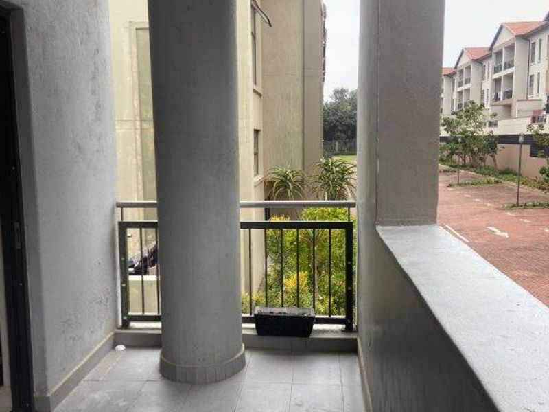 A covered balcony featuring tiled flooring, a metal balustrade, and a structural concrete column, offering an outdoor space with views of the complex grounds.