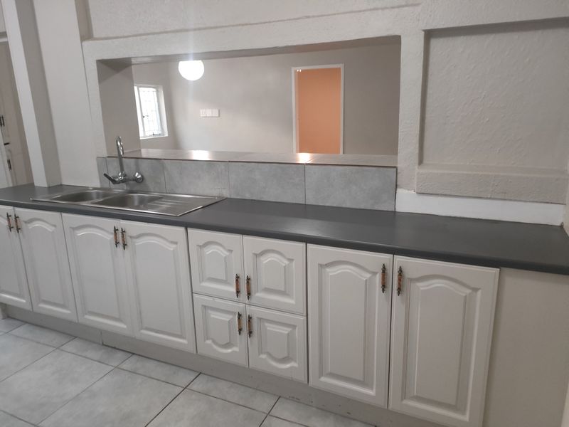 A functional kitchen area featuring white built-in cabinetry, dark laminate countertops, a stainless steel sink, and neutral tiled flooring with a serving hatch opening.