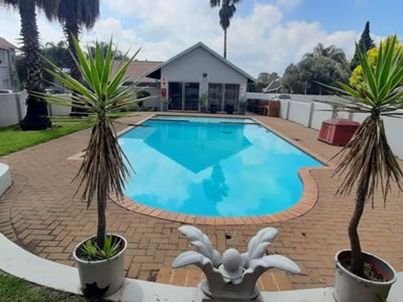 A well-maintained outdoor swimming pool area featuring brick paving, surrounding boundary walls, and established garden landscaping within a townhouse complex.