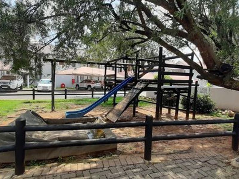 Communal garden area featuring a playground, mature trees, paved pathways, and timber fencing within a townhouse complex.