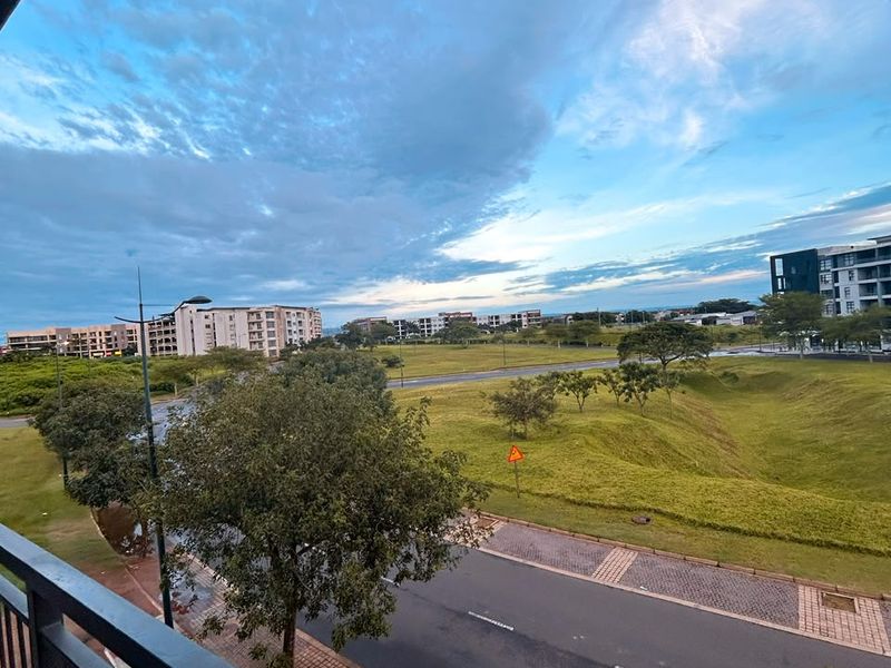An elevated view from a balcony overlooking a landscaped green space, paved roads, and surrounding residential apartment buildings under a cloudy sky.