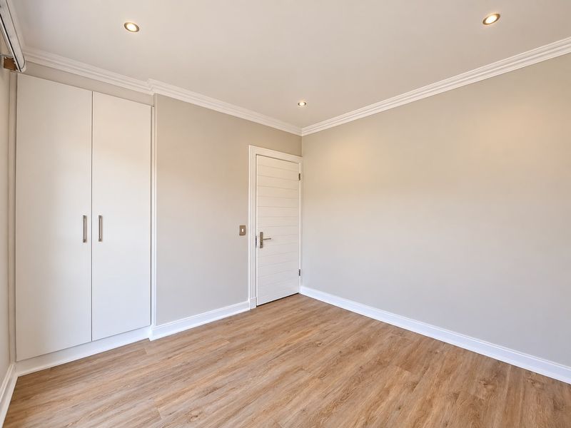 A bright, empty bedroom featuring modern laminate flooring, built-in wardrobes, recessed downlighting, and neutral wall finishes.