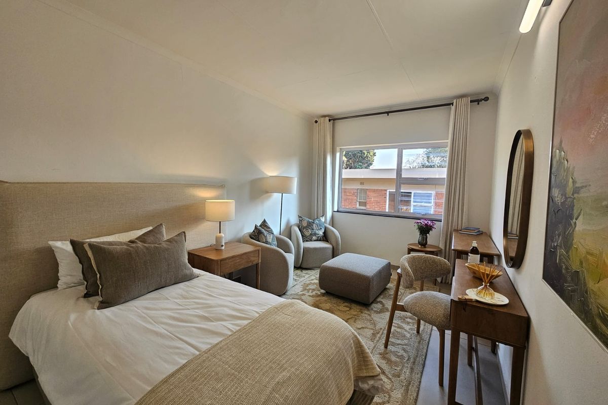 A well-lit, comfortable bedroom featuring neutral tones, tiled flooring, and a large window providing natural light.