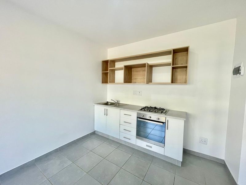 A clean, modern kitchen featuring white cabinetry, built-in oven, gas hob, stainless steel sink, and open wooden shelving on a tiled floor.