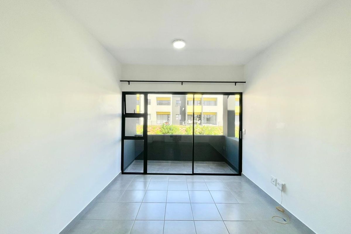 A bright, empty living area featuring neutral tiled flooring and a large sliding glass door that opens onto a balcony.