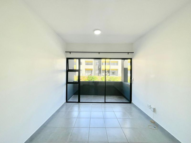 A bright, empty living area featuring neutral tiled flooring and a large sliding glass door that opens onto a balcony.