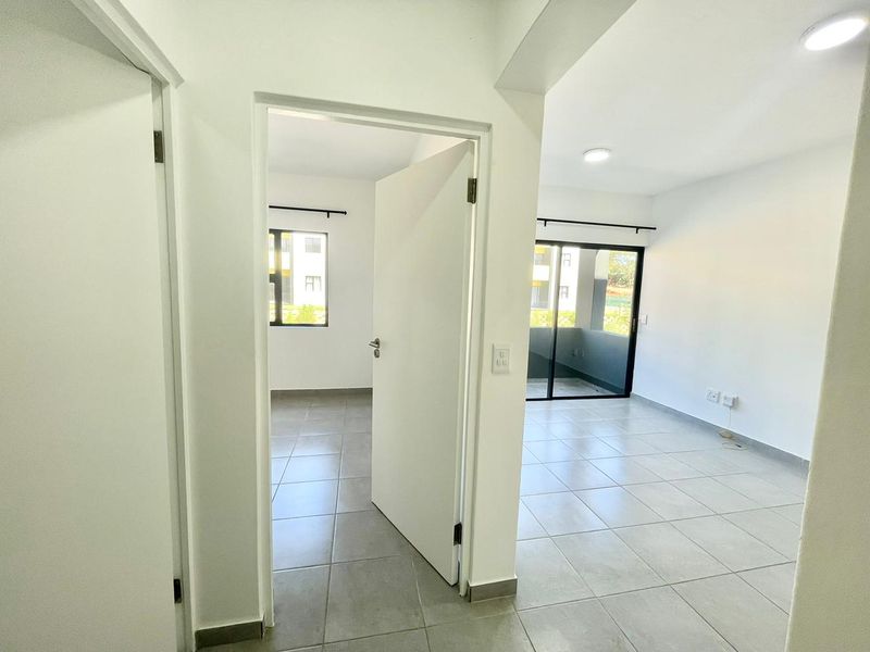 A bright, empty hallway with neutral tiled flooring, leading into an open-plan living area with access to a balcony through sliding glass doors.