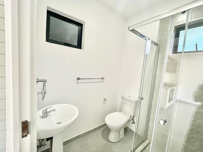 A clean, modern bathroom featuring a pedestal sink, a toilet, and a glass-enclosed shower with neutral floor tiling.