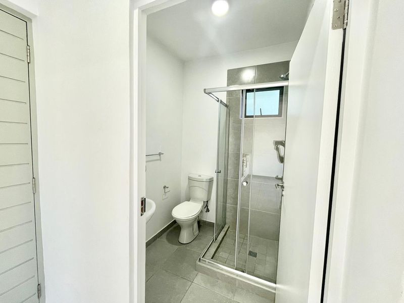 A clean, modern bathroom featuring a glass-enclosed shower, a white toilet, and a wall-mounted basin with neutral grey floor and wall tiling.