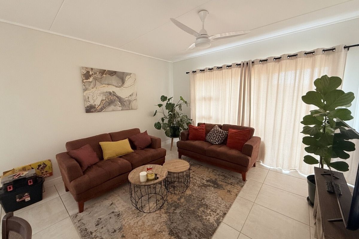 A bright living room featuring neutral tiled flooring, a ceiling fan, and large windows with curtain tracks, providing a comfortable and functional living space.