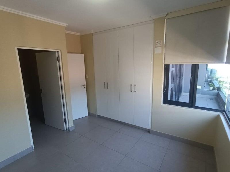 A bright, empty bedroom featuring neutral tiled flooring, built-in white cupboards, and a large window with roller blinds, offering a clean, modern aesthetic.