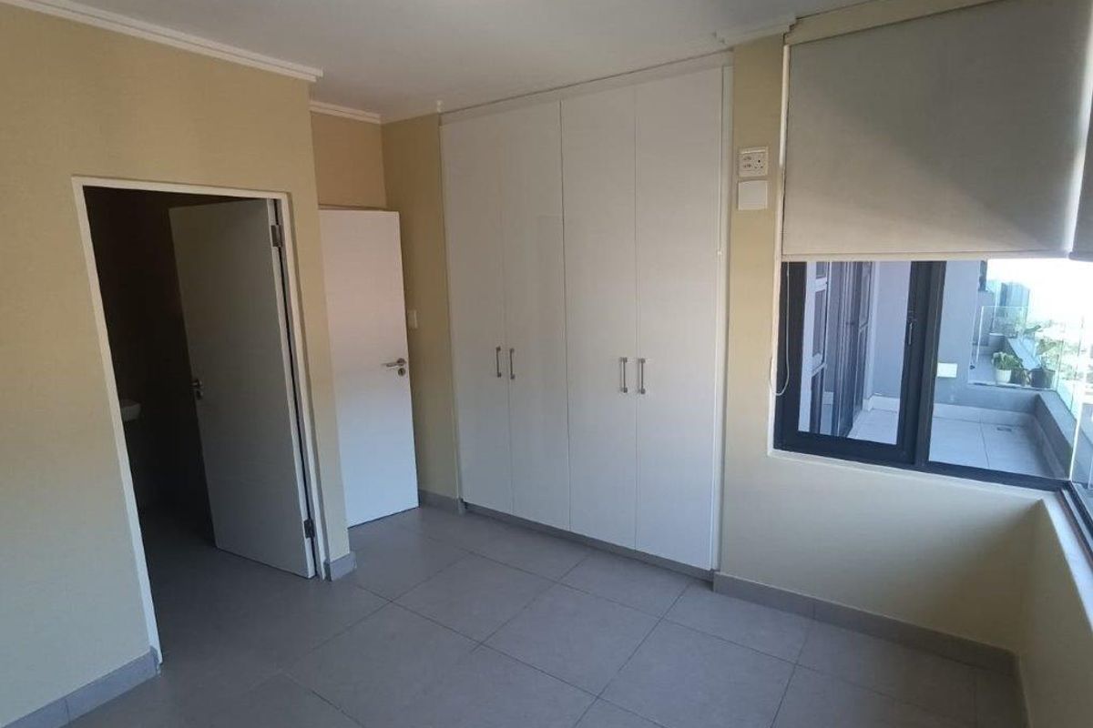 A bright, empty bedroom featuring neutral tiled flooring, built-in white cupboards, and a large window with roller blinds, offering a clean, modern aesthetic.
