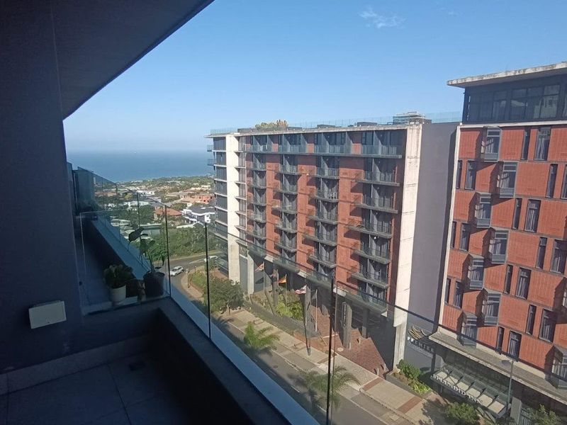 A modern apartment balcony featuring a sleek glass balustrade and tiled flooring, offering an elevated urban view towards the coastline.
