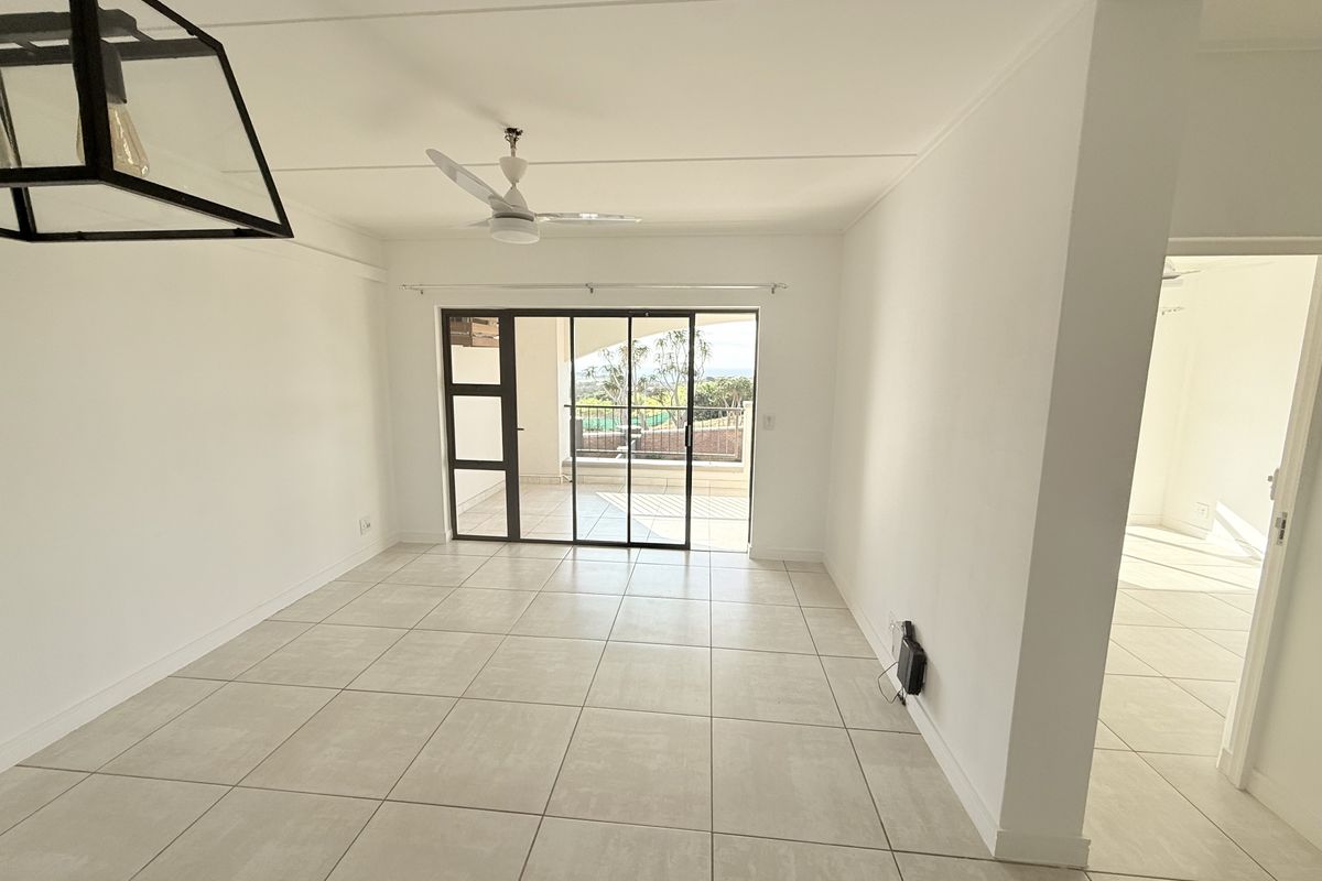 A bright, open-plan living area featuring neutral tiled flooring, a ceiling fan, and large sliding glass doors leading to an outdoor balcony.