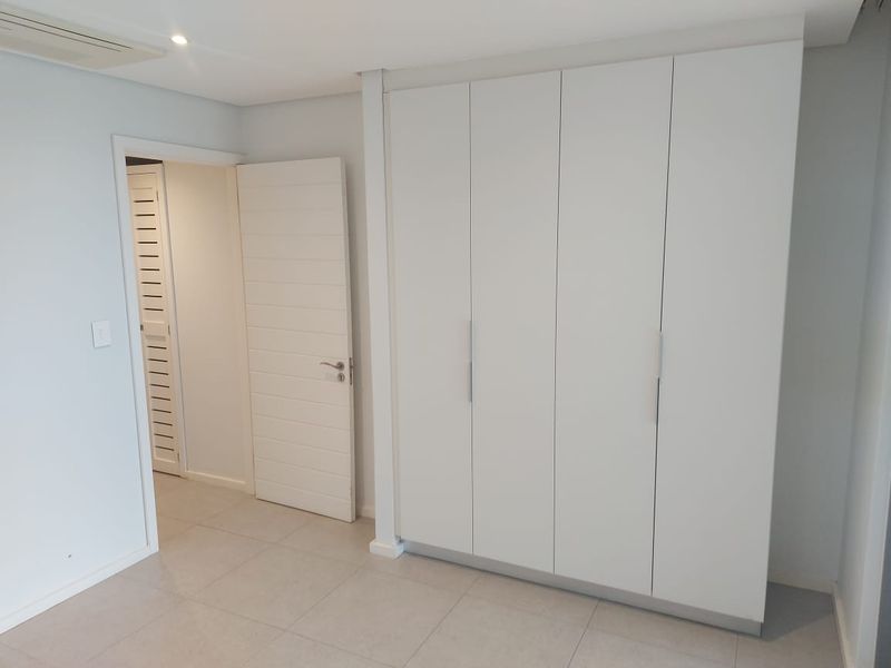 A clean, modern bedroom featuring neutral tiled flooring, recessed lighting, and ample built-in cupboard storage with a minimalist white finish.