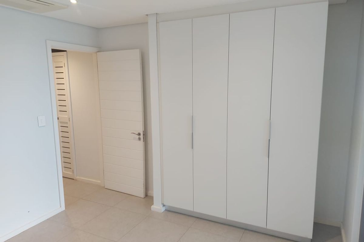 A clean, modern bedroom featuring neutral tiled flooring, recessed lighting, and ample built-in cupboard storage with a minimalist white finish.